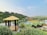 A scenic view of a countryside resort featuring multiple small cabins with green roofs around a central pool. A wooden gazebo with a thatched roof is situated in the forefront, surrounded by lush greenery and flowering plants. The background reveals a hilly landscape with sparse vegetation and several buildings along a ridge.