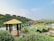 A scenic view of a countryside resort featuring multiple small cabins with green roofs around a central pool. A wooden gazebo with a thatched roof is situated in the forefront, surrounded by lush greenery and flowering plants. The background reveals a hilly landscape with sparse vegetation and several buildings along a ridge.