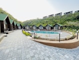 A serene outdoor scene featuring a row of quaint wooden cabins with green roofs lining a paved walkway. In the center lies a round swimming pool surrounded by greenery and outdoor seating. The setting is nestled among lush hills, suggesting a tranquil retreat.