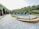 A serene outdoor scene featuring a row of quaint wooden cabins with green roofs lining a paved walkway. In the center lies a round swimming pool surrounded by greenery and outdoor seating. The setting is nestled among lush hills, suggesting a tranquil retreat.