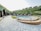 A serene outdoor scene featuring a row of quaint wooden cabins with green roofs lining a paved walkway. In the center lies a round swimming pool surrounded by greenery and outdoor seating. The setting is nestled among lush hills, suggesting a tranquil retreat.