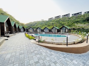 A serene outdoor scene featuring a row of quaint wooden cabins with green roofs lining a paved walkway. In the center lies a round swimming pool surrounded by greenery and outdoor seating. The setting is nestled among lush hills, suggesting a tranquil retreat.