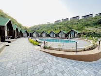 A serene outdoor scene featuring a row of quaint wooden cabins with green roofs lining a paved walkway. In the center lies a round swimming pool surrounded by greenery and outdoor seating. The setting is nestled among lush hills, suggesting a tranquil retreat.