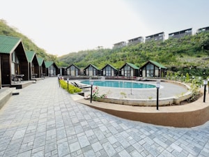 A serene outdoor scene featuring a row of quaint wooden cabins with green roofs lining a paved walkway. In the center lies a round swimming pool surrounded by greenery and outdoor seating. The setting is nestled among lush hills, suggesting a tranquil retreat.