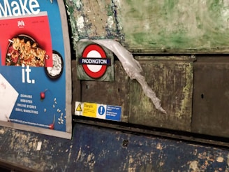 A billboard in a subway station with a colorful advertisement featuring a dish of food. Nearby, the iconic London Underground roundel with 'Paddington' is displayed, partially covered by a translucent plastic sheet. The wall behind appears worn with patches of peeling paint, giving a rustic and gritty look.