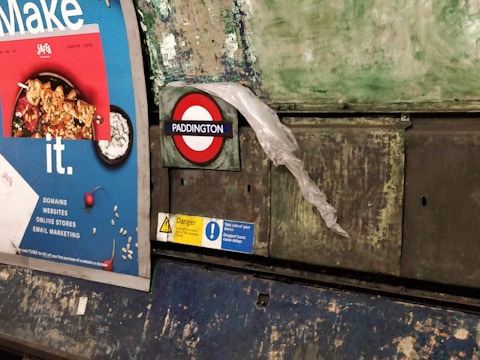 A billboard in a subway station with a colorful advertisement featuring a dish of food. Nearby, the iconic London Underground roundel with 'Paddington' is displayed, partially covered by a translucent plastic sheet. The wall behind appears worn with patches of peeling paint, giving a rustic and gritty look.