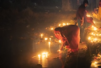 A group of people, some dressed in traditional attire, are gathered near a body of water during nighttime. They are placing lit candles or diyas on the water's surface. The scene is illuminated by the warm glow of these numerous candles, creating a serene and spiritual atmosphere.