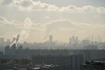 A cityscape featuring numerous high-rise buildings with smoke stacks emitting white smoke. The skyline is hazy, likely due to pollution, and the sky is partly cloudy.