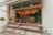 A cozy cafe with an open window bar setup featuring cushioned benches for seating. The interior is decorated with hanging plants and warm glowing lights. Shelves are lined with bottles, and there are various plants on the counter, contributing to a natural and inviting ambiance.