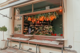 A cozy cafe with an open window bar setup featuring cushioned benches for seating. The interior is decorated with hanging plants and warm glowing lights. Shelves are lined with bottles, and there are various plants on the counter, contributing to a natural and inviting ambiance.
