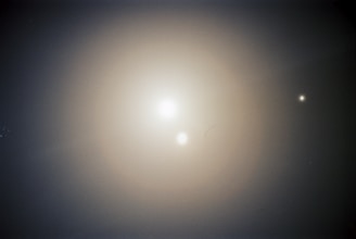 A celestial scene with two bright, glowing orbs against a dark, soft gradient background. The larger orb is centrally positioned, while a smaller one appears in the upper right quadrant. The subtle light scattering adds a mystical and serene quality.
