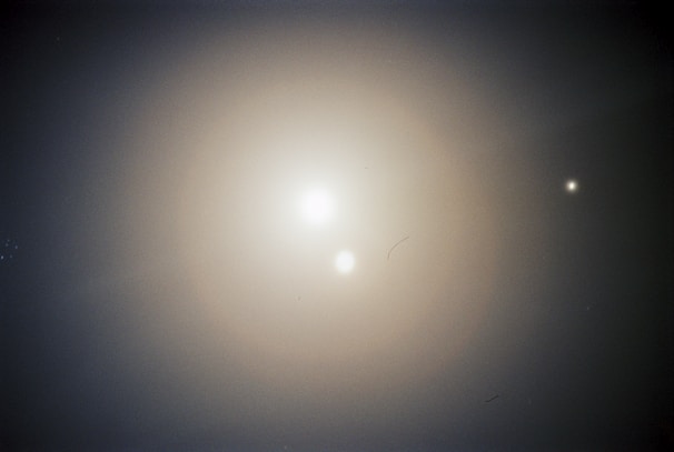 A celestial scene with two bright, glowing orbs against a dark, soft gradient background. The larger orb is centrally positioned, while a smaller one appears in the upper right quadrant. The subtle light scattering adds a mystical and serene quality.