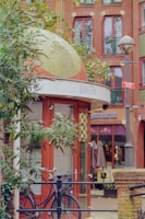 A quaint urban scene with a small, dome-shaped bakery building featuring a mosaic roof and red-framed glass doors. A bicycle is parked in front, next to some green foliage. The background includes a multi-story building with arched windows and a small outdoor café area with umbrellas and chairs.