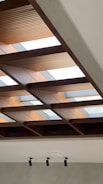 A modern ceiling design featuring a grid of wooden beams with integrated skylights, allowing natural light to enter the room. The wooden beams are contrasted by a smooth grey wall below, equipped with multiple cylindrical ceiling lights.