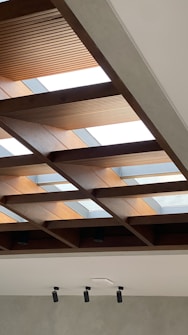 A modern ceiling design featuring a grid of wooden beams with integrated skylights, allowing natural light to enter the room. The wooden beams are contrasted by a smooth grey wall below, equipped with multiple cylindrical ceiling lights.