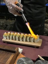 A person wearing gloves uses a small torch to sear the top of a sushi roll placed on a wooden board. The sushi roll is garnished with toppings and sauces. Various items including a towel, chopsticks, and a wooden box are visible on the table.