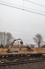 An excavator is actively moving a pile of gravel near a train track, with freight containers in the background. Bare trees and a misty sky create a somber atmosphere.
