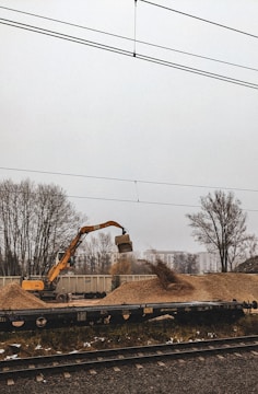 An excavator is actively moving a pile of gravel near a train track, with freight containers in the background. Bare trees and a misty sky create a somber atmosphere.