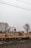 An excavator is actively moving a pile of gravel near a train track, with freight containers in the background. Bare trees and a misty sky create a somber atmosphere.