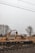 An excavator is actively moving a pile of gravel near a train track, with freight containers in the background. Bare trees and a misty sky create a somber atmosphere.