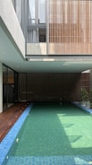 A modern indoor swimming pool is surrounded by a minimalist architectural structure. The pool is long with clear water and blue tiles. Wooden paneling and large windows contribute to an open and airy atmosphere. Sunlight filters through, casting shadows on the water.