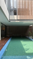 A modern indoor swimming pool is surrounded by a minimalist architectural structure. The pool is long with clear water and blue tiles. Wooden paneling and large windows contribute to an open and airy atmosphere. Sunlight filters through, casting shadows on the water.