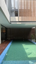 A modern indoor swimming pool is surrounded by a minimalist architectural structure. The pool is long with clear water and blue tiles. Wooden paneling and large windows contribute to an open and airy atmosphere. Sunlight filters through, casting shadows on the water.