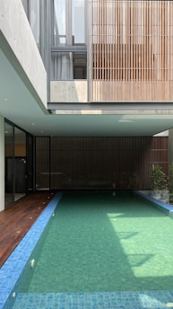A modern indoor swimming pool is surrounded by a minimalist architectural structure. The pool is long with clear water and blue tiles. Wooden paneling and large windows contribute to an open and airy atmosphere. Sunlight filters through, casting shadows on the water.