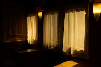 Warm, ambient lighting filters through sheer curtains, casting a soft glow across the dark wooden interior. The room exudes a cozy and serene atmosphere, enhanced by the interplay of light and shadow on the walls and surfaces.