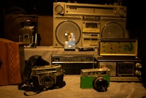 A collection of vintage items including radios, cameras, and a clock. The central focus is on an old boombox with large speakers. Beside it, there are classic film cameras and a brightly colored analog radio. A decorative clock with a panda design is also visible, adding a nostalgic feel to the scene.