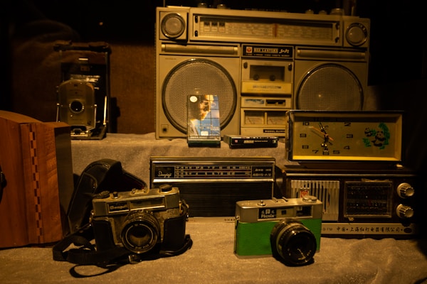 A collection of vintage items including radios, cameras, and a clock. The central focus is on an old boombox with large speakers. Beside it, there are classic film cameras and a brightly colored analog radio. A decorative clock with a panda design is also visible, adding a nostalgic feel to the scene.