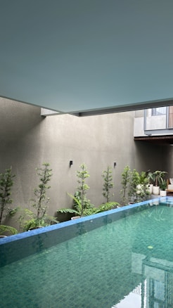 An indoor swimming pool with clear, blue-green water is surrounded by small, potted plants along the perimeter on a light-colored wall. The ceiling is a smooth, muted blue-gray. The poolside features a modern architectural element, including minimalistic design and clean lines.