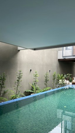 An indoor swimming pool with clear, blue-green water is surrounded by small, potted plants along the perimeter on a light-colored wall. The ceiling is a smooth, muted blue-gray. The poolside features a modern architectural element, including minimalistic design and clean lines.
