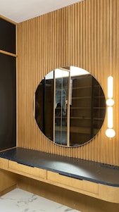 A modern interior features a circular mirror mounted on a vertical wooden panel with a slatted design. Beneath the mirror is a dark countertop with a subtle marbled pattern, supported by a light wooden structure with drawers. Two vertical light fixtures are positioned beside the mirror, providing soft illumination.