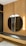 A modern interior features a circular mirror mounted on a vertical wooden panel with a slatted design. Beneath the mirror is a dark countertop with a subtle marbled pattern, supported by a light wooden structure with drawers. Two vertical light fixtures are positioned beside the mirror, providing soft illumination.