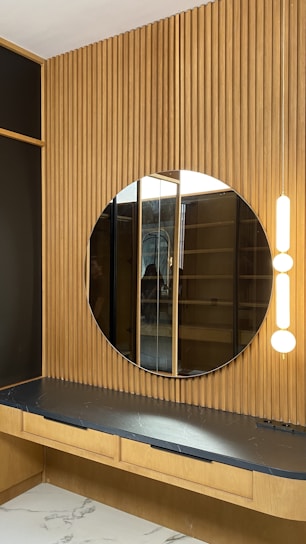 A modern interior features a circular mirror mounted on a vertical wooden panel with a slatted design. Beneath the mirror is a dark countertop with a subtle marbled pattern, supported by a light wooden structure with drawers. Two vertical light fixtures are positioned beside the mirror, providing soft illumination.