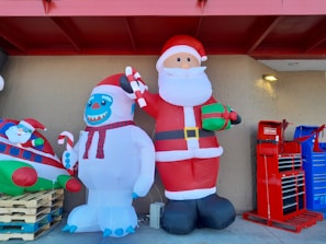 Two large inflatable decorations are displayed outside a building. One is a Santa Claus holding a gift and a candy cane. Next to Santa is a snowman-like creature with blue facial features, also holding a candy cane. To the left, part of another inflatable featuring Santa inside a plane is visible. On the right, there are red and blue tool cabinets.