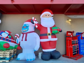 Two large inflatable decorations are displayed outside a building. One is a Santa Claus holding a gift and a candy cane. Next to Santa is a snowman-like creature with blue facial features, also holding a candy cane. To the left, part of another inflatable featuring Santa inside a plane is visible. On the right, there are red and blue tool cabinets.