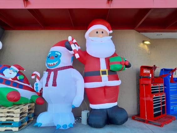 Two large inflatable decorations are displayed outside a building. One is a Santa Claus holding a gift and a candy cane. Next to Santa is a snowman-like creature with blue facial features, also holding a candy cane. To the left, part of another inflatable featuring Santa inside a plane is visible. On the right, there are red and blue tool cabinets.