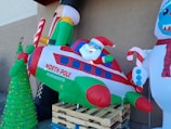 An inflatable display featuring Santa Claus in a festive airplane labeled 'North Pole Airways.' A nutcracker holds a candy cane on the left, while a snowman with a scarf stands to the right. A decorated Christmas tree with multicolored lights is positioned in front.