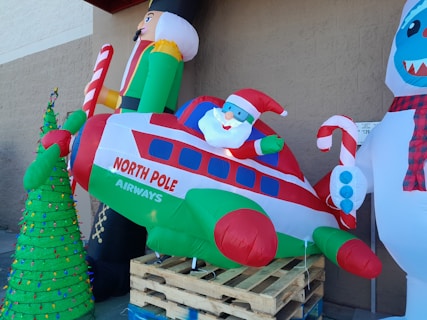 An inflatable display featuring Santa Claus in a festive airplane labeled 'North Pole Airways.' A nutcracker holds a candy cane on the left, while a snowman with a scarf stands to the right. A decorated Christmas tree with multicolored lights is positioned in front.