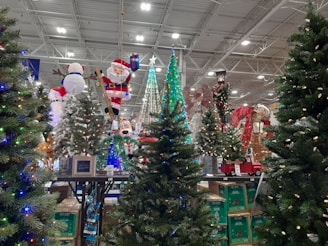 A festive display featuring a variety of artificial Christmas trees adorned with colorful lights and decorations. Among the decorations is an inflatable Santa Claus holding a present and a snowman. In the background, reindeer and candy cane decorations can be seen, alongside a model of a red truck carrying a small tree.
