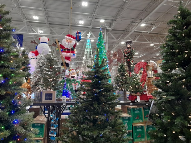 A festive display featuring a variety of artificial Christmas trees adorned with colorful lights and decorations. Among the decorations is an inflatable Santa Claus holding a present and a snowman. In the background, reindeer and candy cane decorations can be seen, alongside a model of a red truck carrying a small tree.