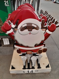 A stack of metal or plastic decorative signs featuring a cartoon Santa Claus with a red hat and reindeer antlers. The signs are arranged on a wooden pallet and displayed in a retail setting. The price tag on the pallet indicates the cost.