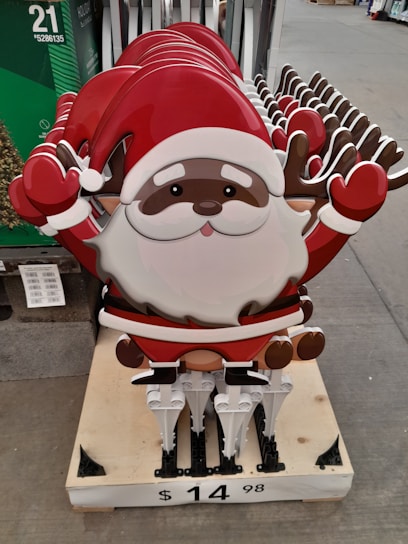 A stack of metal or plastic decorative signs featuring a cartoon Santa Claus with a red hat and reindeer antlers. The signs are arranged on a wooden pallet and displayed in a retail setting. The price tag on the pallet indicates the cost.