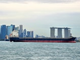 A large cargo ship is sailing on a body of water with a cityscape in the background, featuring modern skyscrapers and a distinctive three-towered hotel with a rooftop terrace.