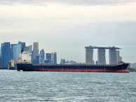 A large cargo ship is sailing on a body of water with a cityscape in the background, featuring modern skyscrapers and a distinctive three-towered hotel with a rooftop terrace.