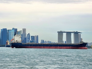 A large cargo ship is sailing on a body of water with a cityscape in the background, featuring modern skyscrapers and a distinctive three-towered hotel with a rooftop terrace.