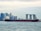 A large cargo ship is sailing on a body of water with a cityscape in the background, featuring modern skyscrapers and a distinctive three-towered hotel with a rooftop terrace.