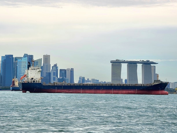 A large cargo ship is sailing on a body of water with a cityscape in the background, featuring modern skyscrapers and a distinctive three-towered hotel with a rooftop terrace.